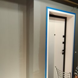 Valeriy - Świeżo pomalowana szara ściana z widocznymi śladami po szpachlowaniu, zabezpieczone taśmą malarską ościeżnice drzwi wejściowych z białą skrzydłem i czarnymi okuciami, krzesło metalowe w tle.