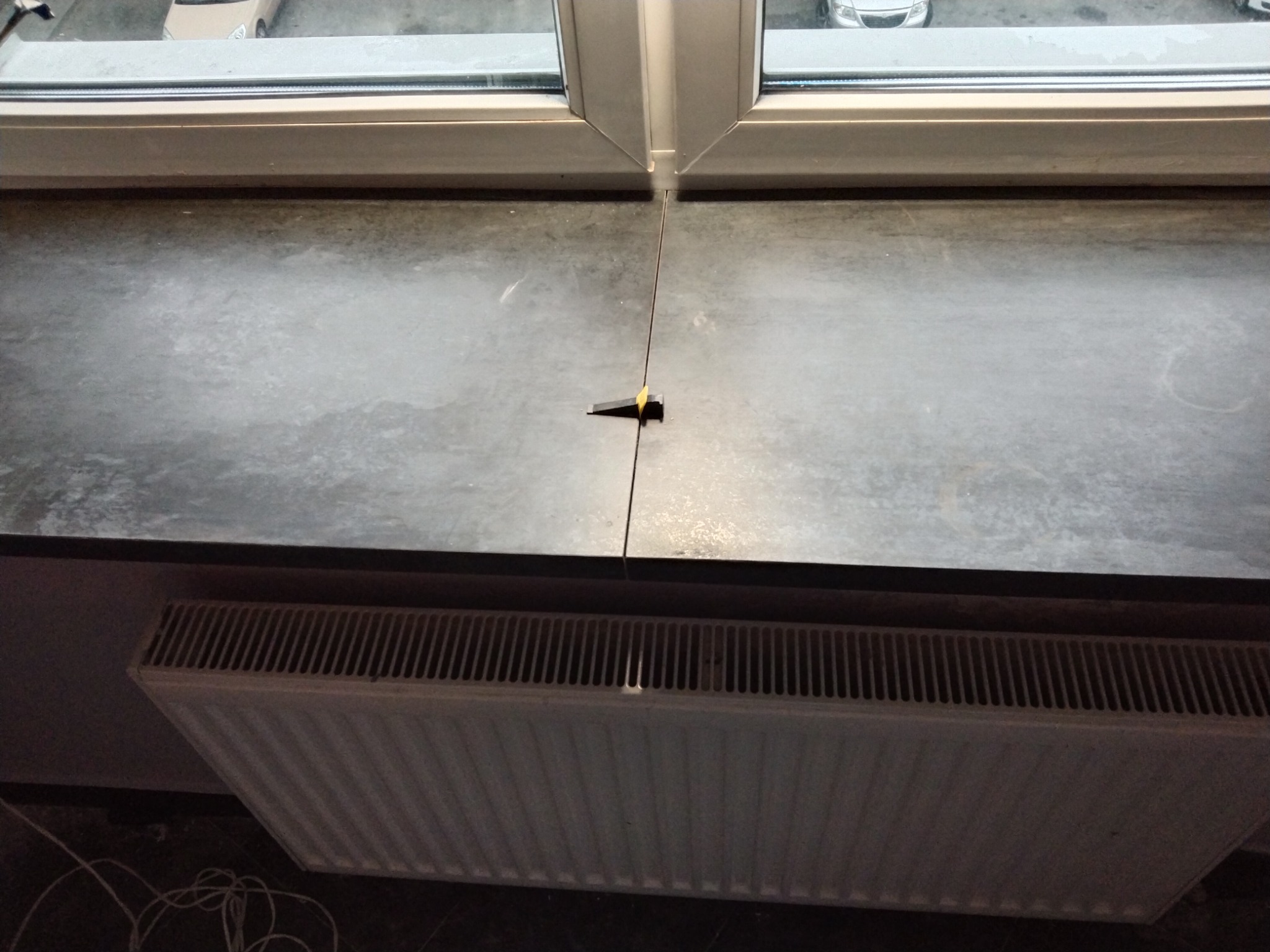 Detal wstawiania płytek na parapecie okiennym z zastosowaniem klina dystansowego, widoczny grzejnik pod parapetem i fragment okna z widokiem na parking.