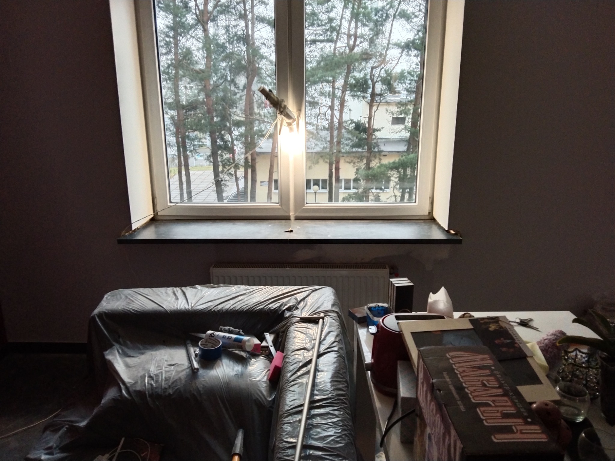 Pokój w trakcie przygotowań do prac remontowych: meble przykryte folią ochronną, widoczne narzędzia i materiały budowlane, okno z widokiem na drzewa, prowizoryczne oświetlenie.
