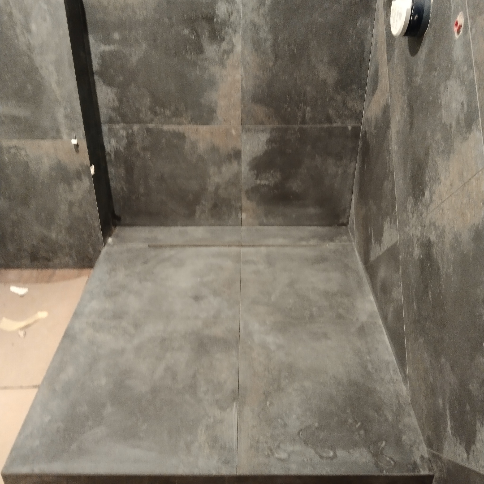 Szare, matowe płytki ceramiczne ułożone w kabinie prysznicowej typu walk-in, widoczny odpływ liniowy i fragment instalacji wodnej na ścianie.