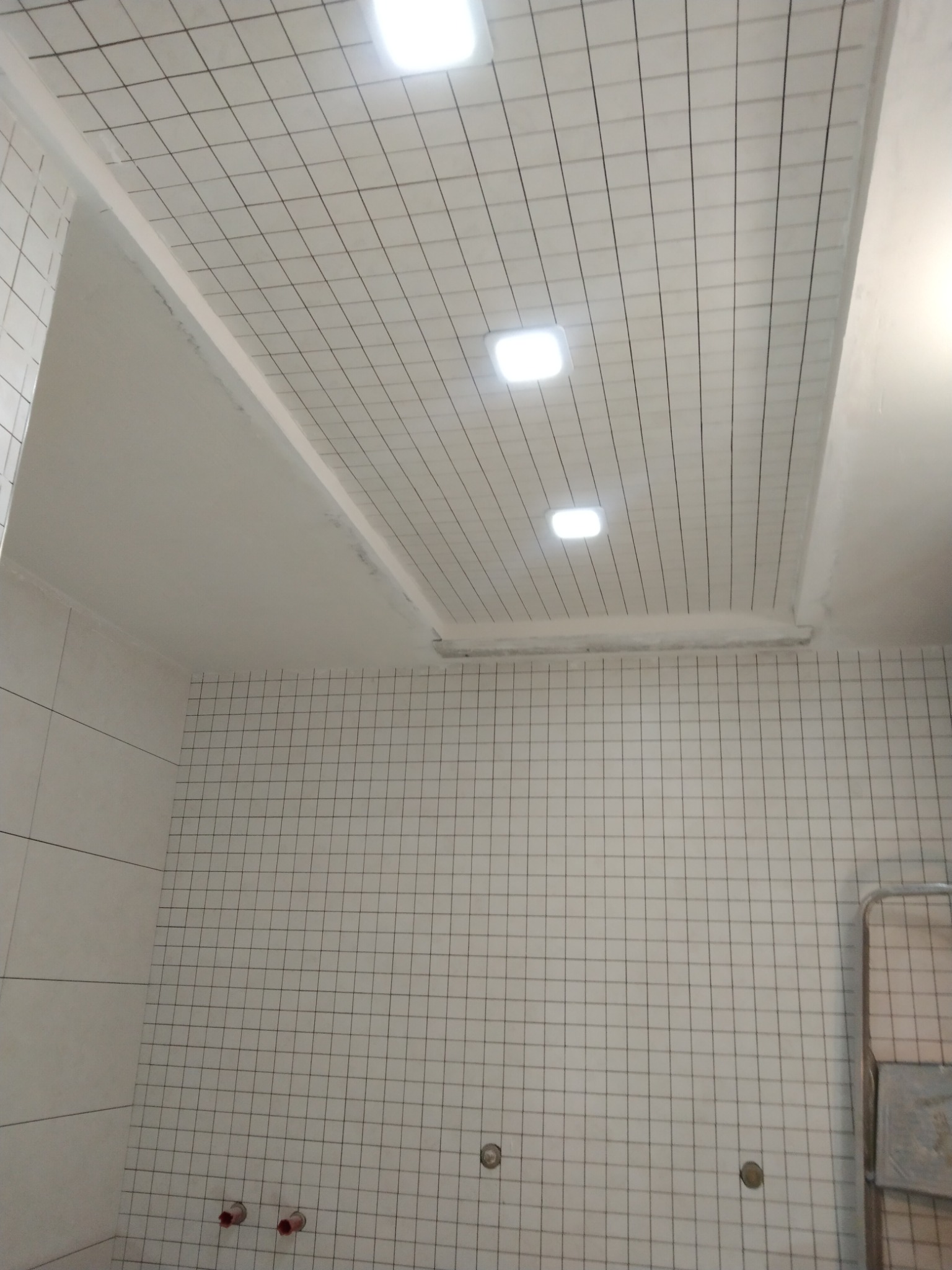 Biały sufit z kwadratową siatką i wbudowanymi punktami świetlnymi, dopasowany do ściany z białych płytek z czarną siatką fug, widoczne instalacje hydrauliczne przy podłodze.