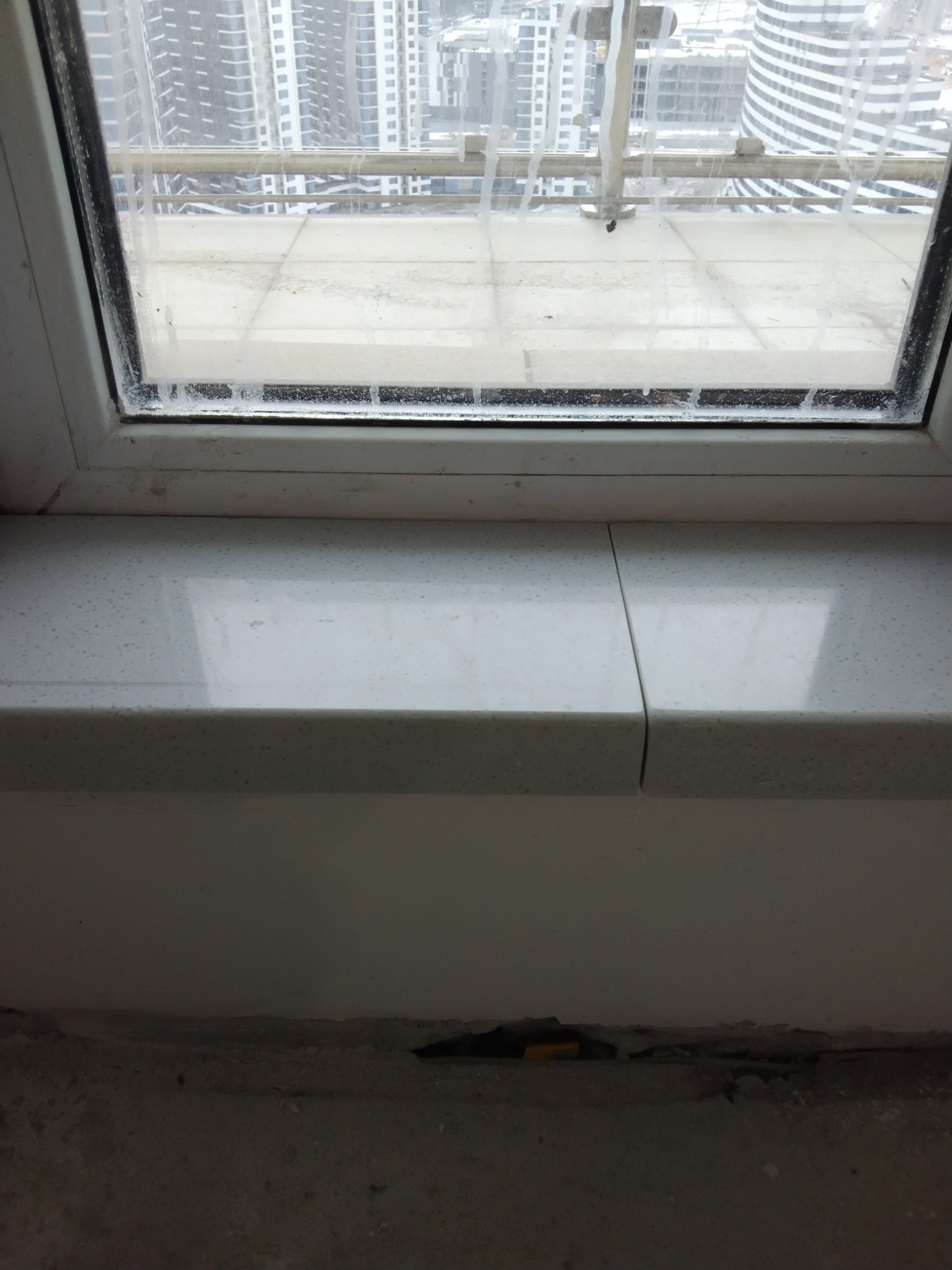 Widok z okna na panoramę miasta z zamontowanym, białym parapetem z widocznym łączeniem dwóch elementów, zacieki na szybie i niedokończone wykończenie pod parapetem.