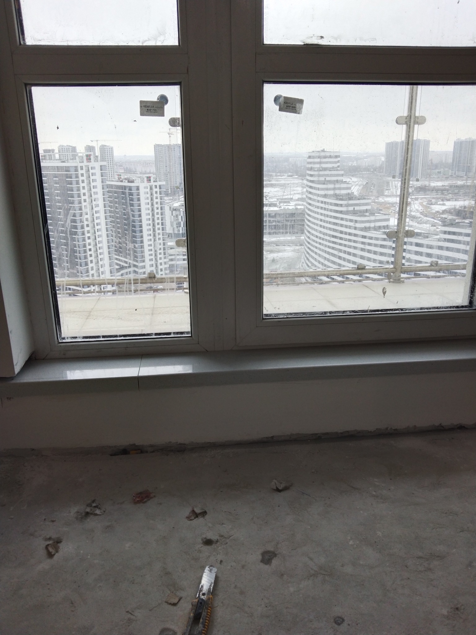 Widok z okna na panoramę miasta z wysokiego piętra w trakcie remontu, na podłodze resztki materiałów i nóż monterski.