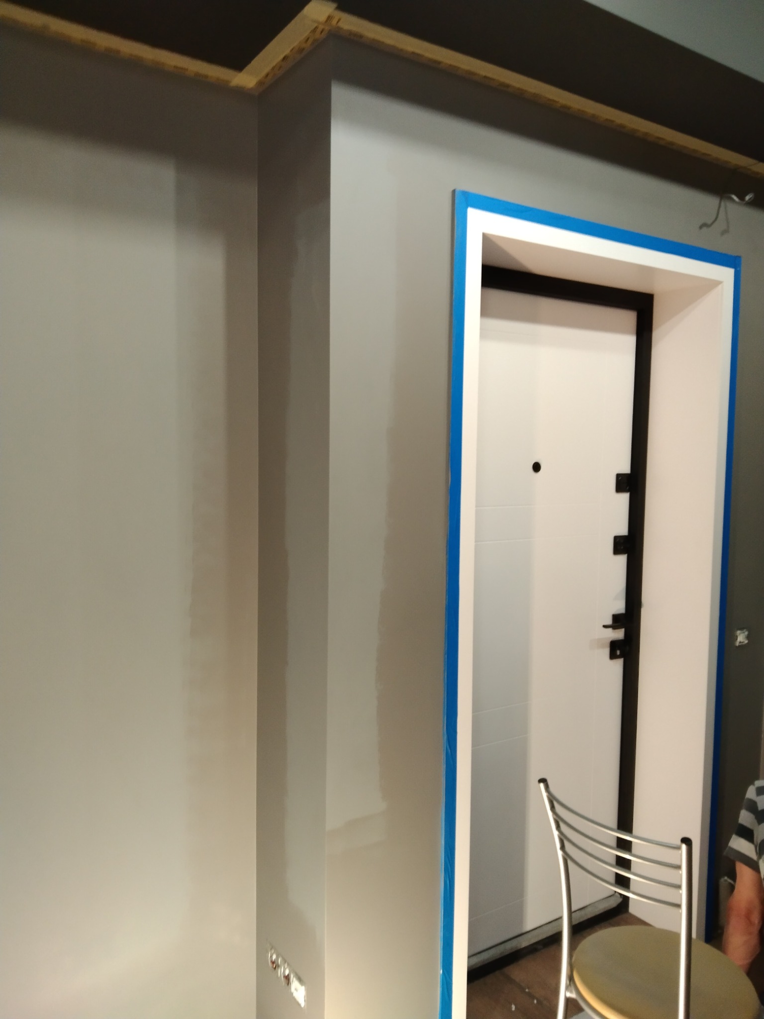 Świeżo pomalowana szara ściana z widocznymi śladami po szpachlowaniu, zabezpieczone taśmą malarską ościeżnice drzwi wejściowych z białą skrzydłem i czarnymi okuciami, krzesło metalowe w tle.