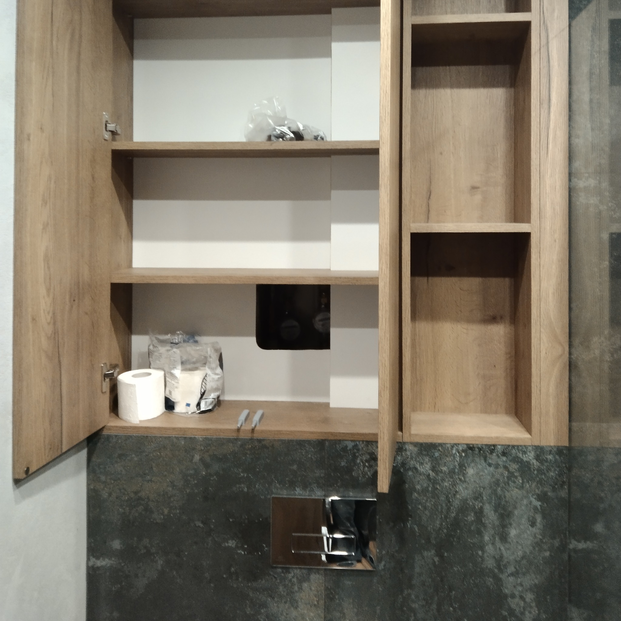 Otwarta szafka łazienkowa z drewnopodobnymi frontami i półkami, widoczne akcesoria łazienkowe i instalacja za szafką, na tle ściany wykończonej ciemną płytką imitującą kamień.