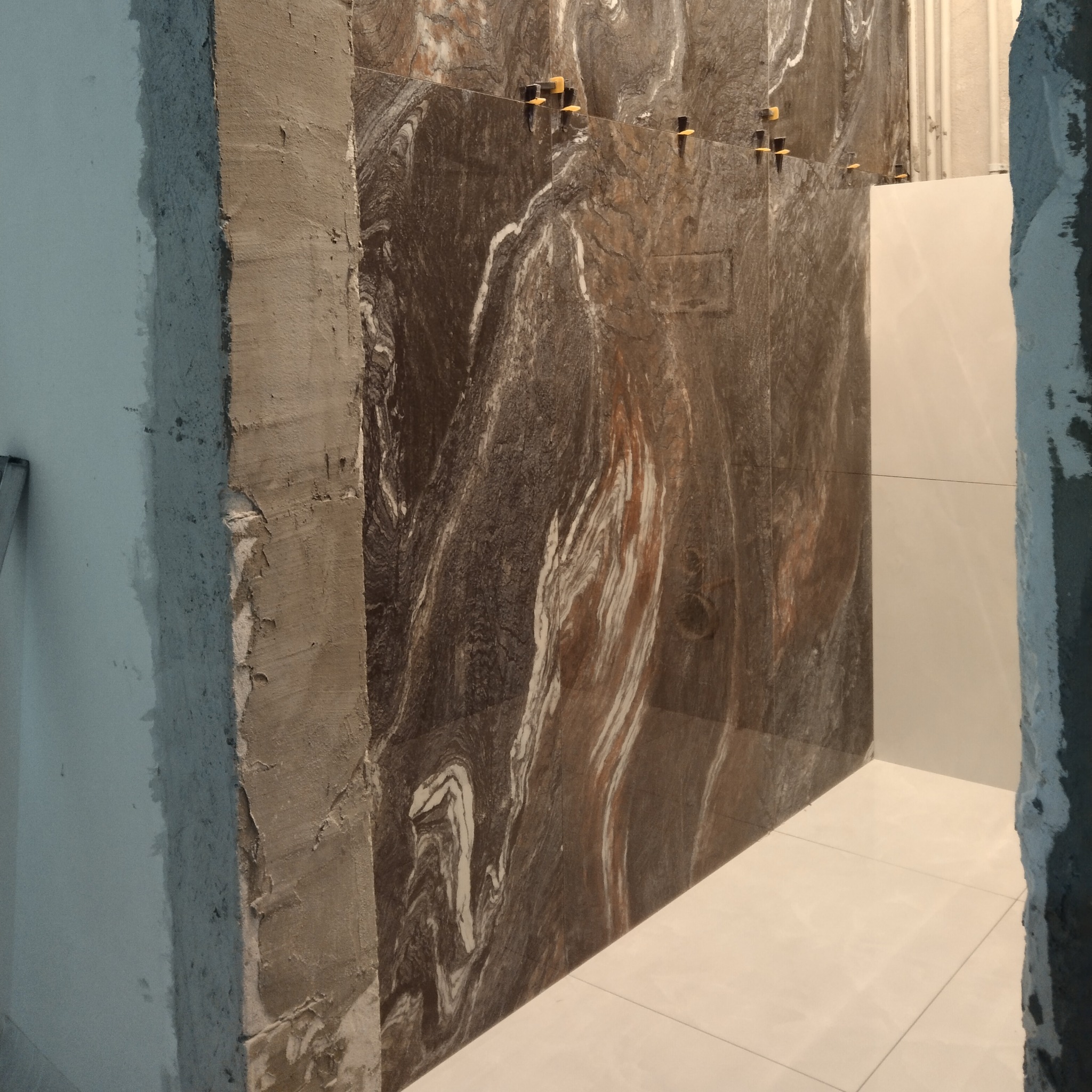 Montaż płytek imitujących marmur na ścianie, widoczne krzyżyki dystansowe, surowa ściana w tle.