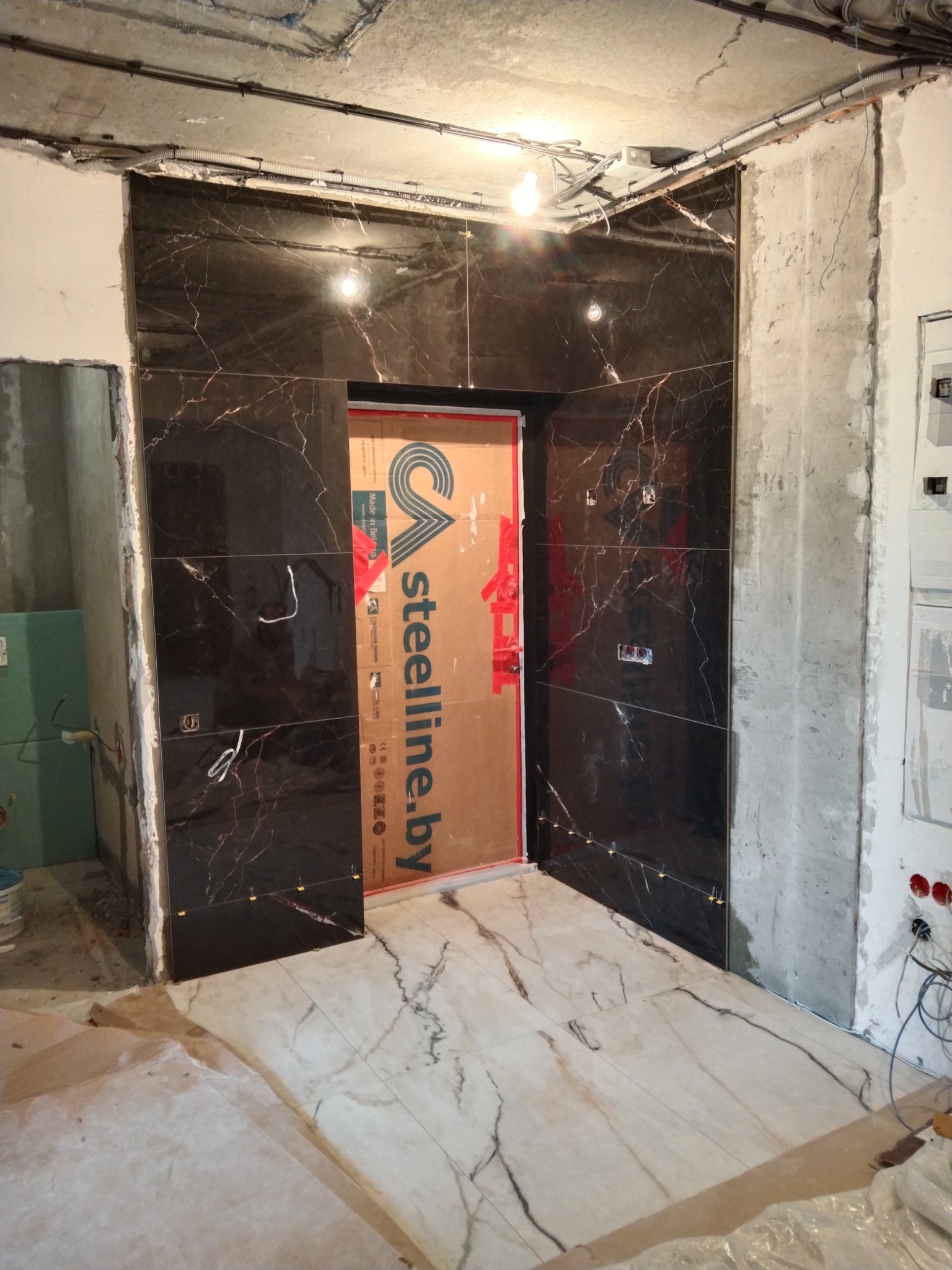 Montaż płytek imitujących marmur na ścianie i podłodze w surowym wnętrzu, widoczne kable i puszki elektryczne, zabezpieczone drzwi kartonem.