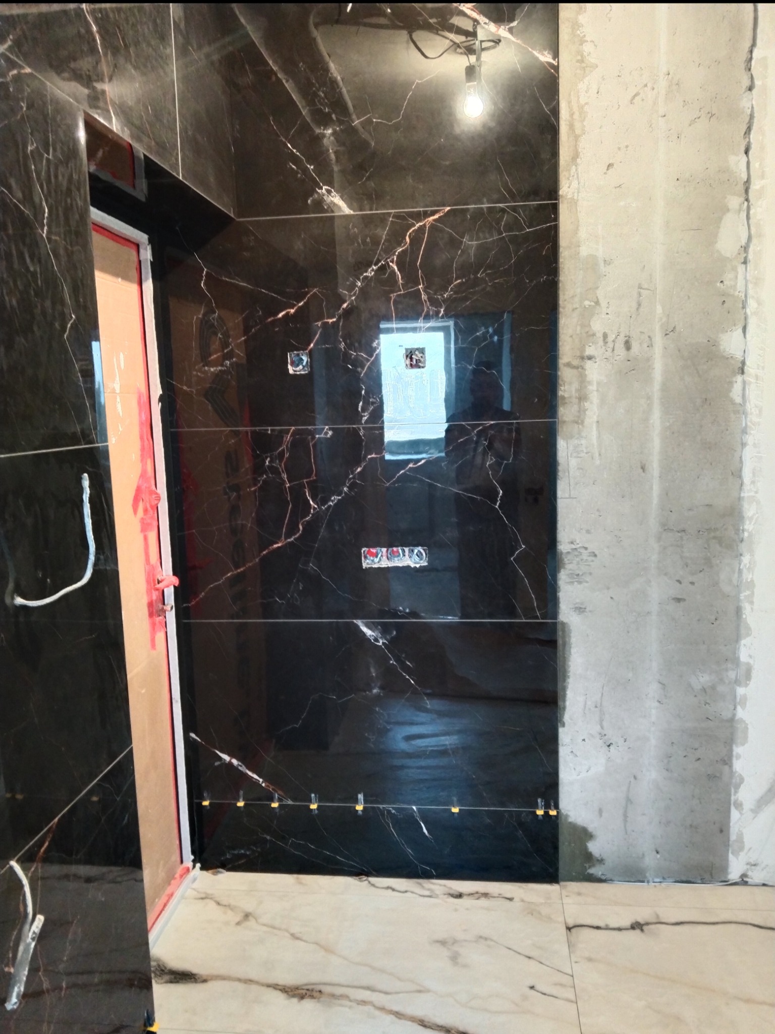 Montaż czarnych płytek imitujących marmur na ścianie łazienki, widoczne krzyżyki dystansowe, surowa ściana obok, instalacja podtynkowa.