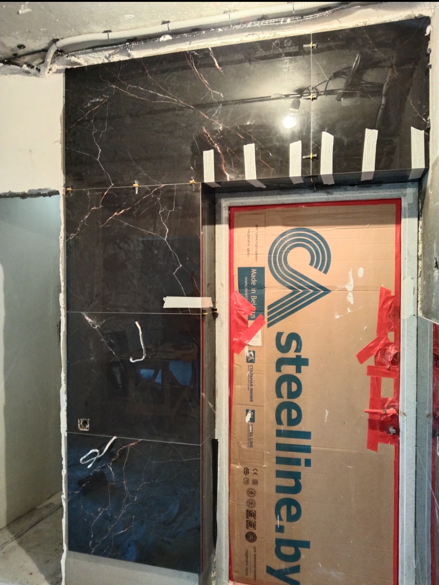 Montaż czarnych płytek imitujących marmur na ścianie, zabezpieczone taśmą malarską, widoczne krzyżyki dystansowe, drzwi zabezpieczone kartonem z napisem steelline.by, instalacja elektryczna...