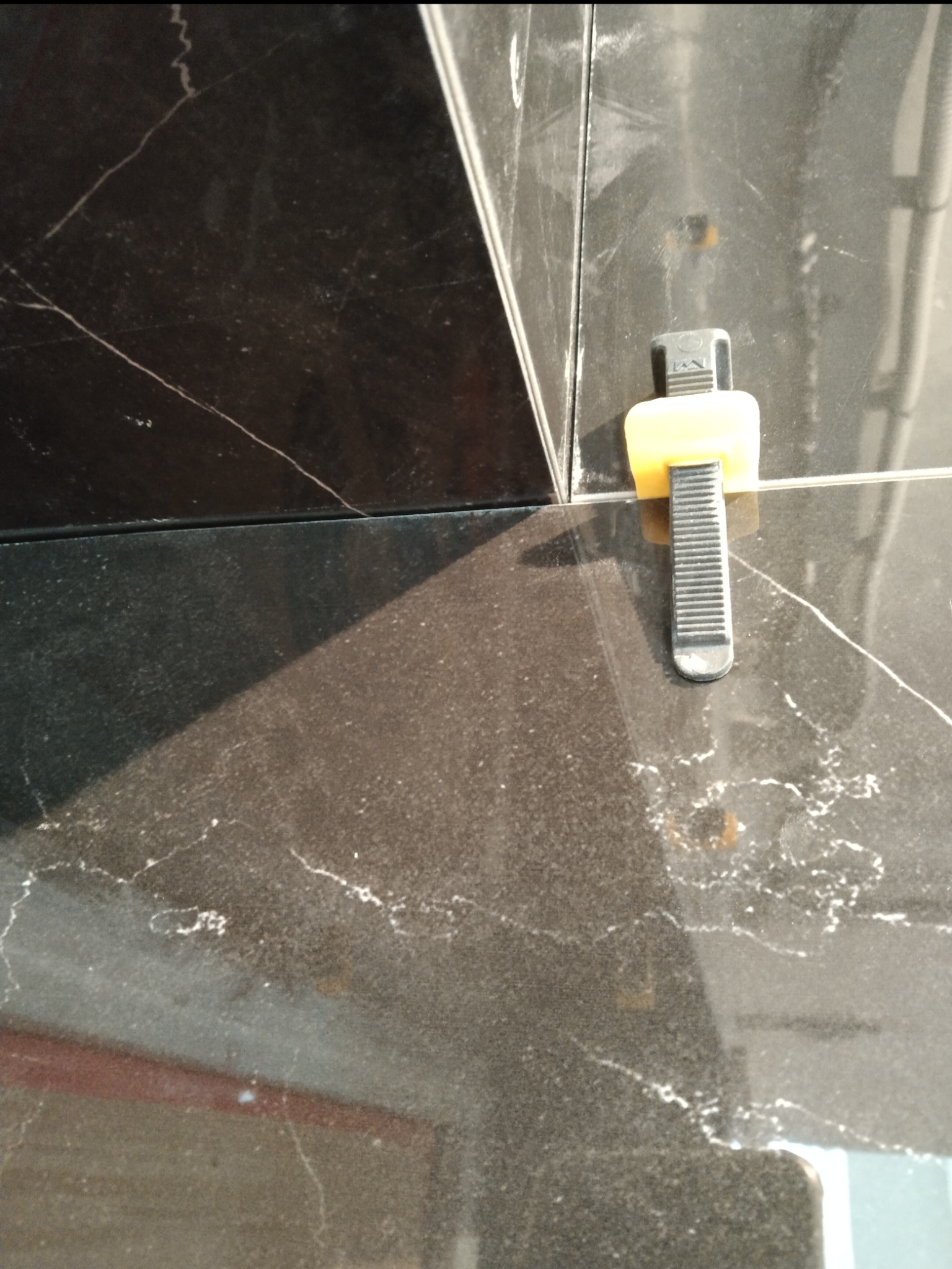 Fragment układania czarnych płytek imitujących marmur z użyciem klipsów poziomujących, widoczne drobne zabrudzenia na powierzchni.