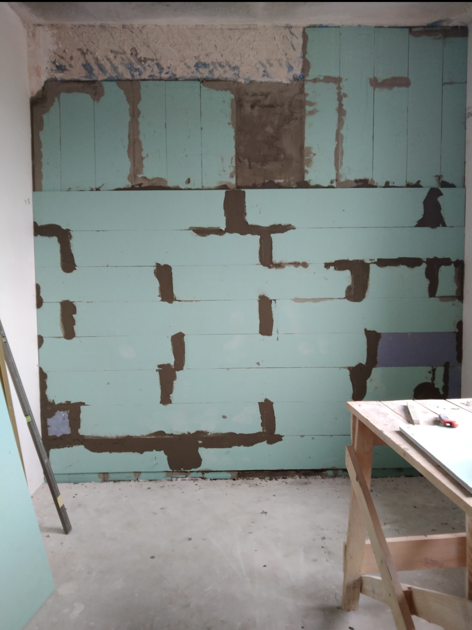 Ściana z płyt styropianowych w trakcie montażu, widoczne ślady kleju montażowego między płytami, fragmenty starego tynku na górze, ustawiona poziomica i stół roboczy w pomieszczeniu.