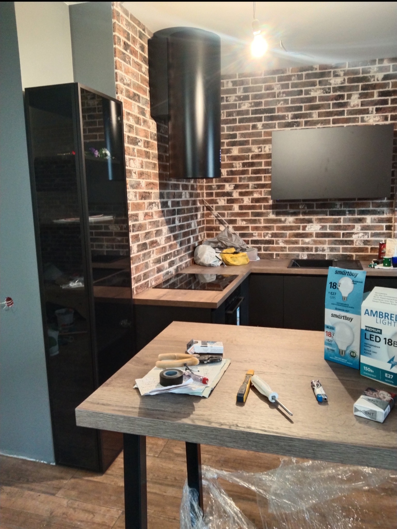 Kuchnia w trakcie prac w Warszawie: widoczna ściana z cegły, czarny okap, czarne szafki kuchenne, narzędzia i materiały budowlane na blacie, w tle czarna witryna.