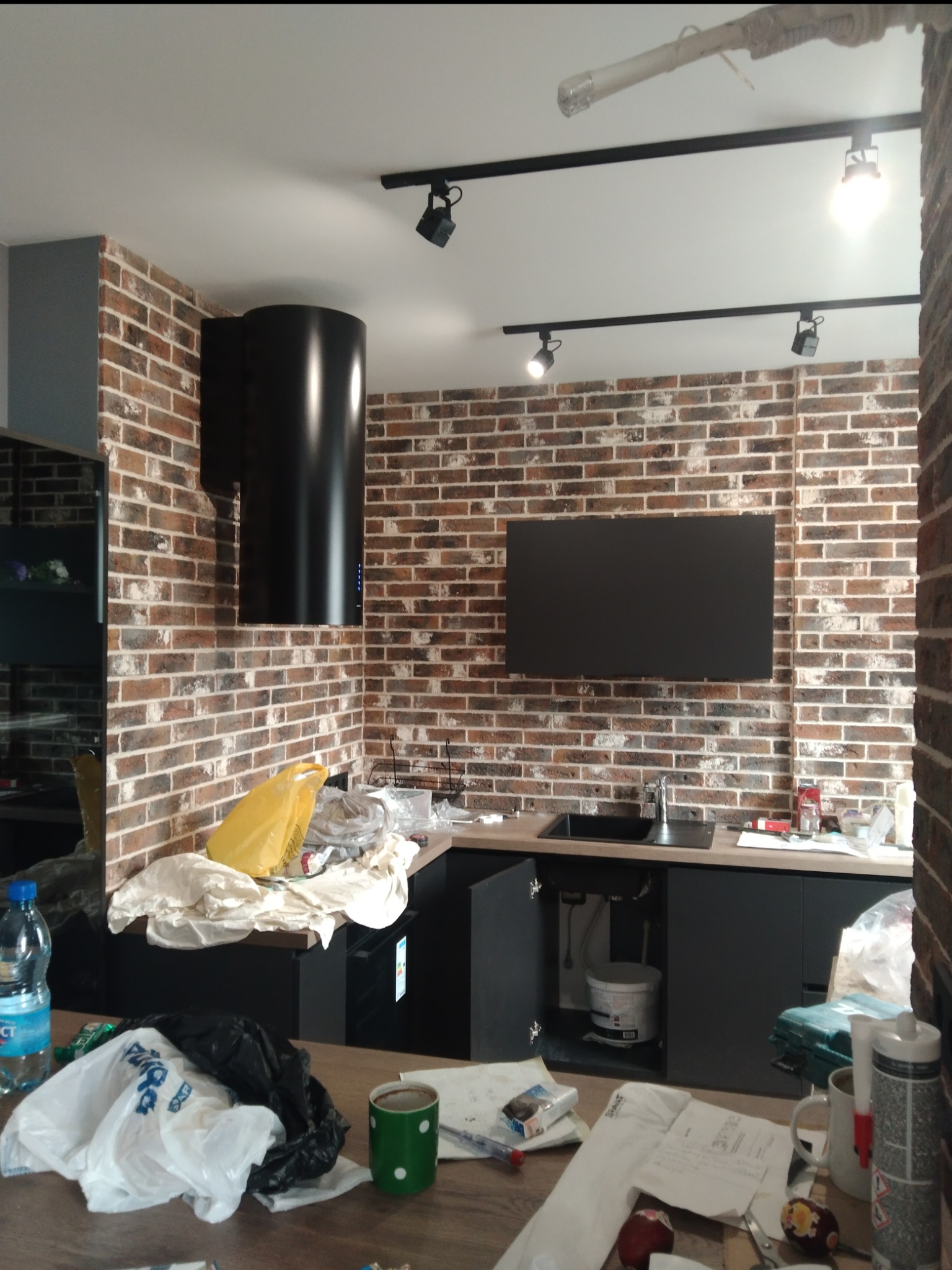 Wnętrze kuchni w trakcie remontu: ściana z cegły, czarne szafki, okap i lampy sufitowe, widoczne narzędzia i materiały budowlane, bałagan na blacie.