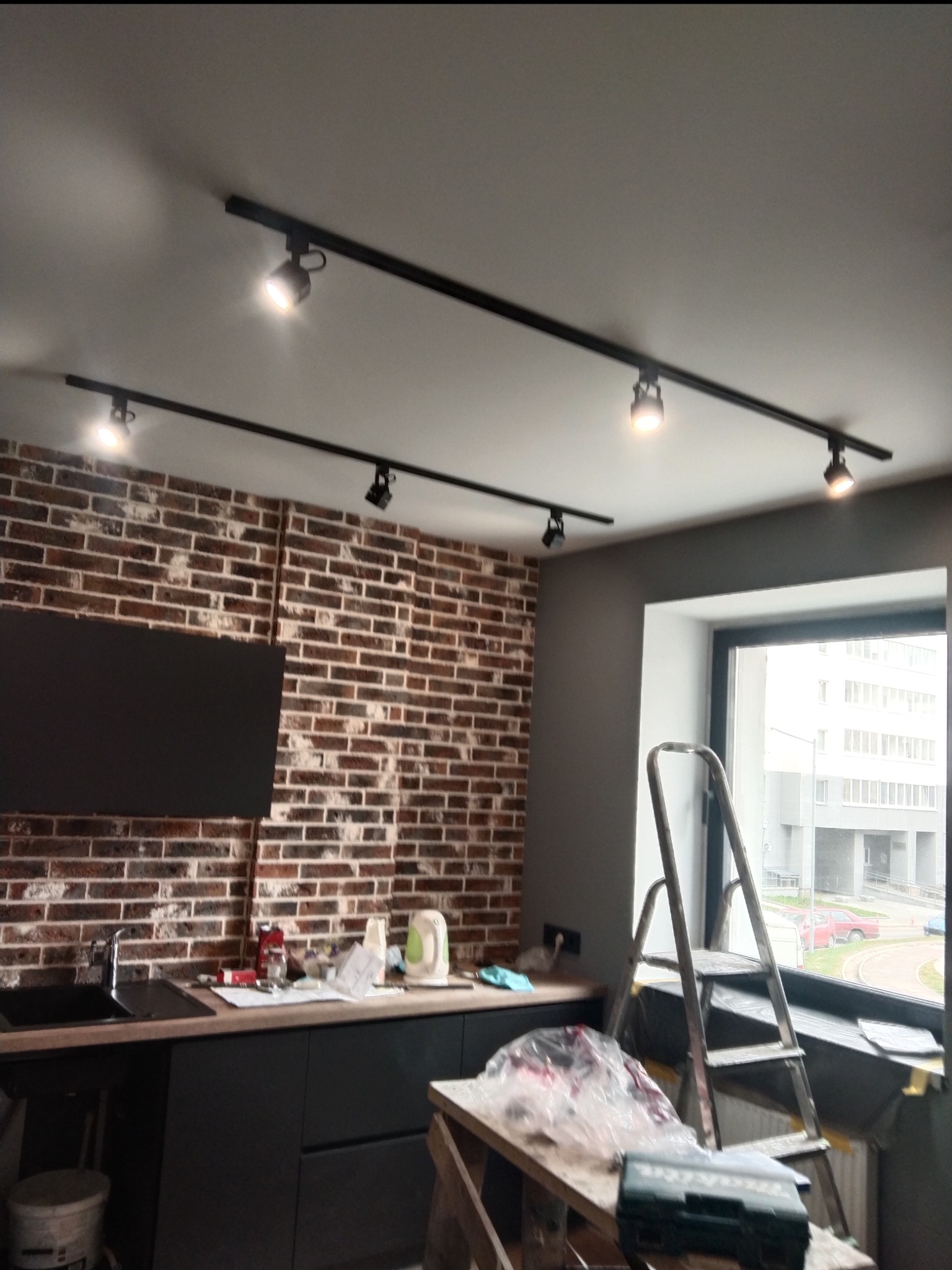 Montaż czarnych szynoprzewodów z reflektorami w kuchni z ceglaną ścianą, widoczna drabina i narzędzia.