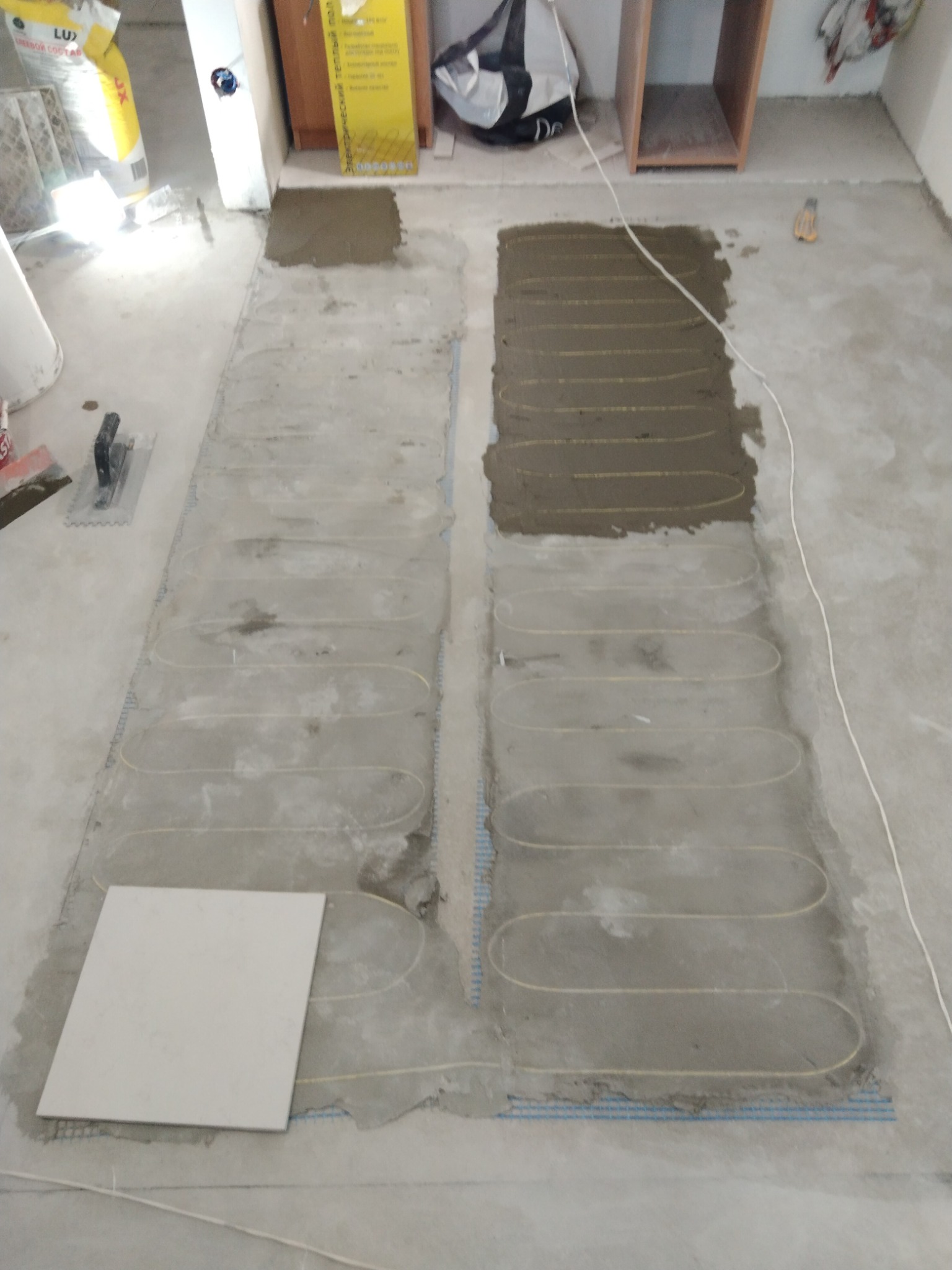 Montaż ogrzewania podłogowego: widoczne kable grzewcze zatopione w wylewce, częściowo pokryte warstwą zaprawy, obok leży płytka gotowa do położenia.