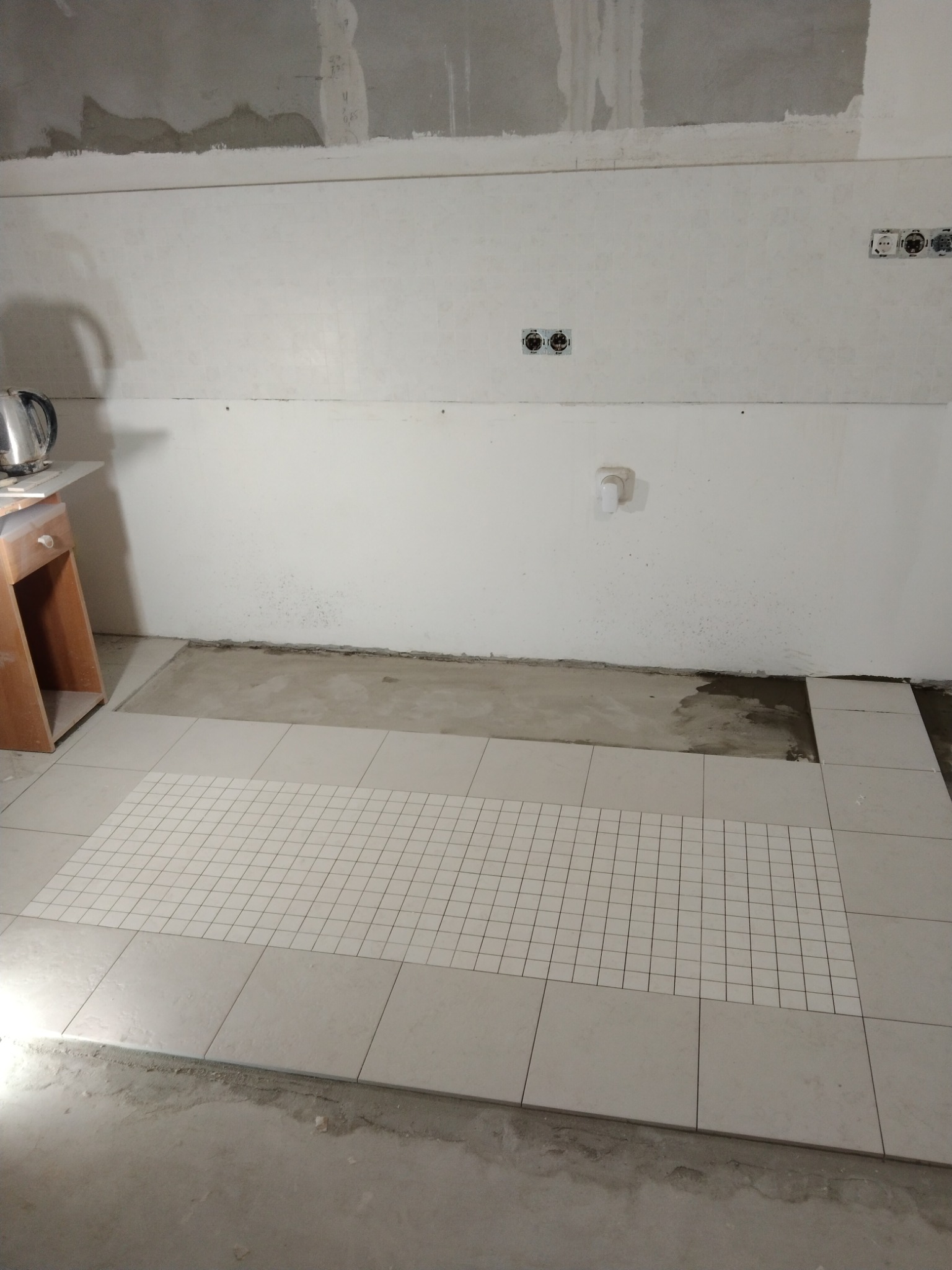 Fragment podłogi w trakcie układania płytek, widoczny fragment siatki montażowej, ściany w stanie surowym, gniazdka elektryczne zamontowane, czajnik na stole w tle.