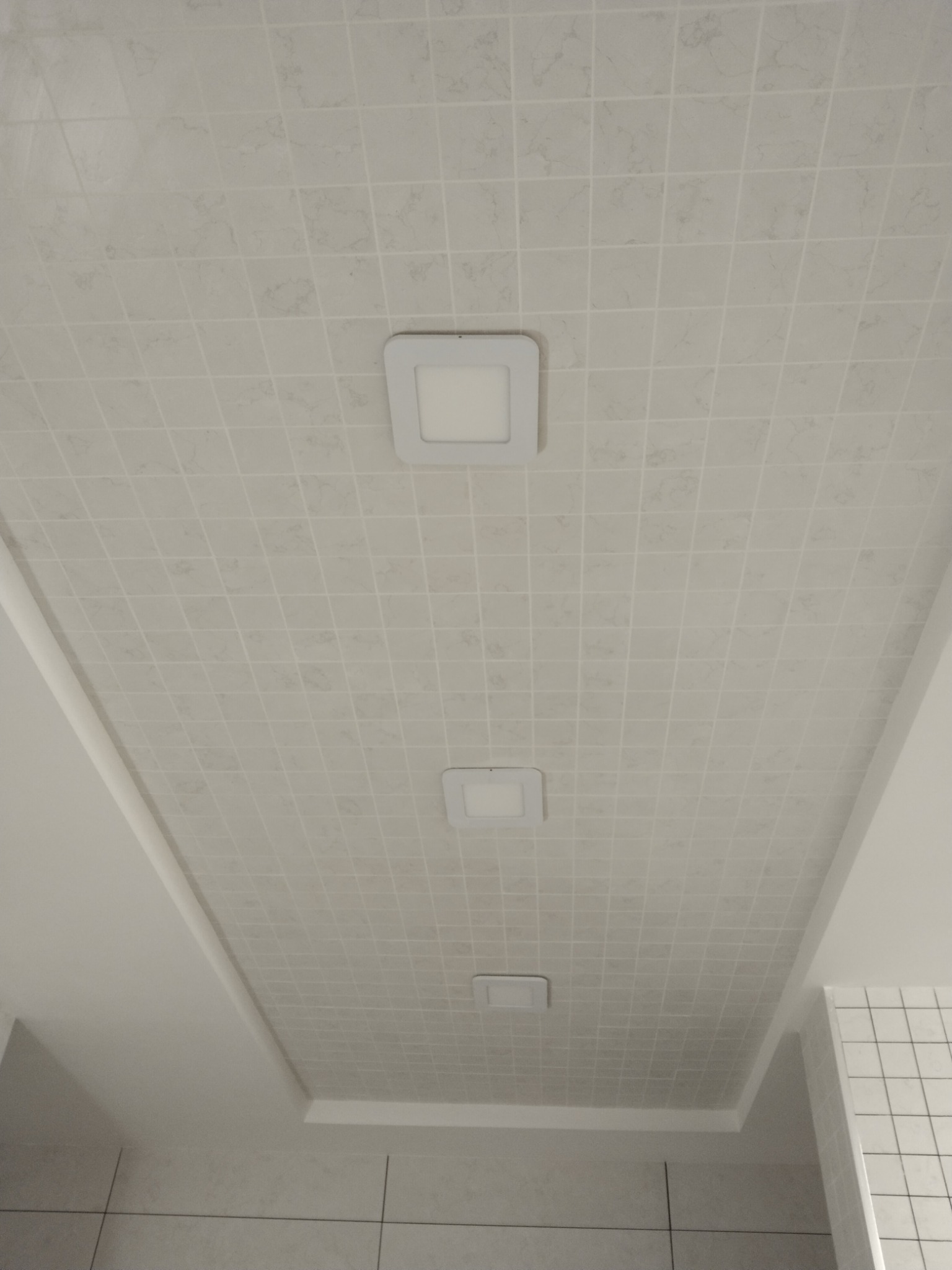 Biały sufit z kwadratowymi panelami i trzema wpuszczonymi lampami LED o kwadratowym kształcie, widok z dołu.