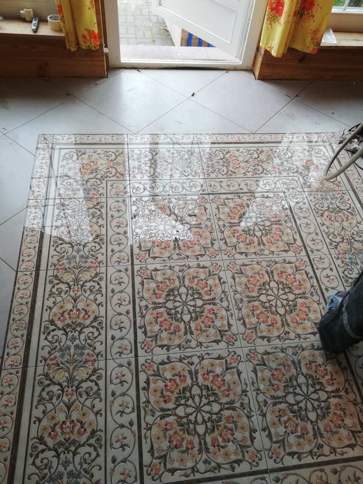 Ułożone płytki podłogowe z motywem kwiatowym, widoczne fragmenty białych fug i otwarte drzwi balkonowe w tle.