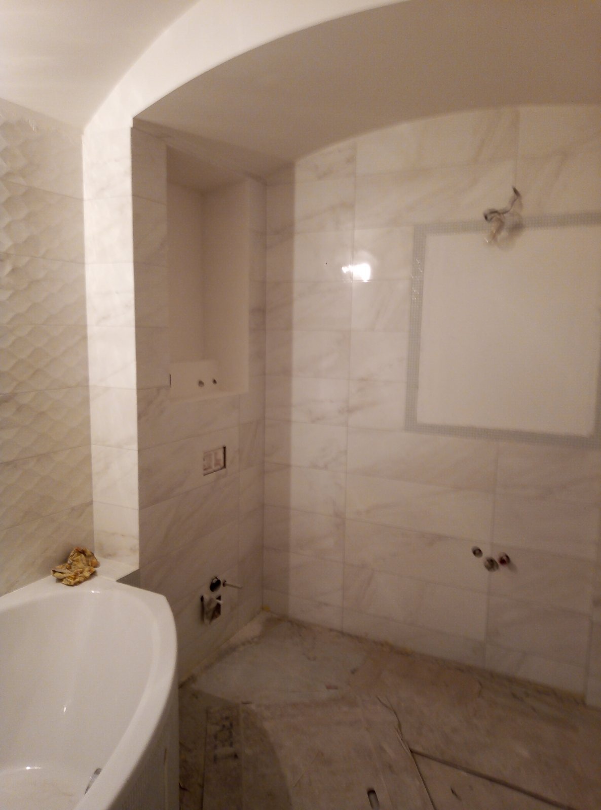 Wnętrze łazienki w trakcie remontu, widoczne białe marmurowe płytki na ścianach, przygotowane miejsce na lustro, fragment wanny i niedokończona podłoga.