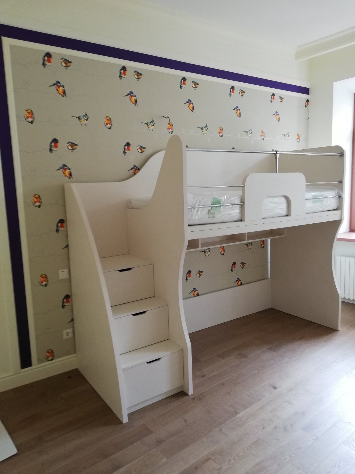 Białe łóżko piętrowe z drabinką z szufladami w pokoju dziecięcym, ściana pokryta tapetą z motywem kolorowych ptaków, widoczne listwy przypodłogowe i fragment okna z grzejnikiem.