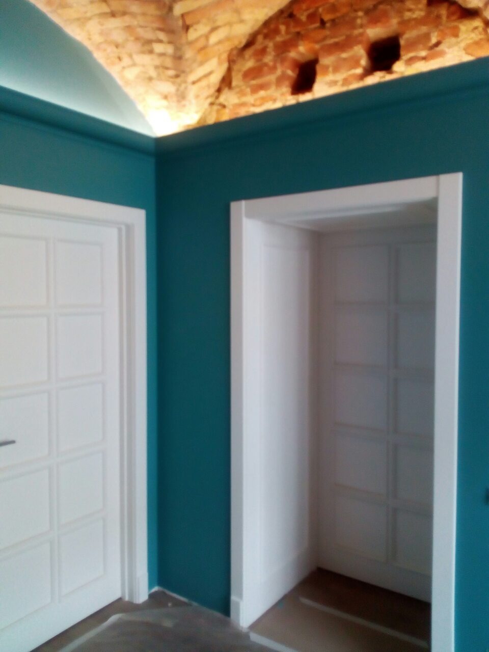Turkusowe ściany z dwoma białymi drzwiami, jedno wejście jest otwarte, sufit z cegły z ukrytym oświetleniem LED.