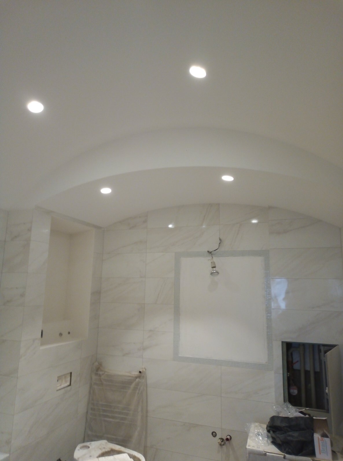 Łazienka w trakcie remontu z zaokrąglonym sufitem podwieszanym z oświetleniem punktowym, marmurowe płytki na ścianach, przygotowane miejsce na lustro z dekoracyjną ramką, widoczne elementy...