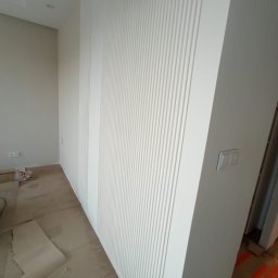 4domy - Biała ściana z dekoracyjnymi panelami pionowymi w trakcie montażu, widoczne gniazdko elektryczne, zabezpieczona podłoga i elementy konstrukcyjne w tle.