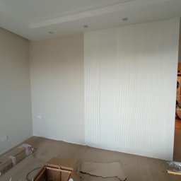 4domy - Pokój po odświeżeniu, z beżowymi ścianami, białą ścianą z pionowymi panelami dekoracyjnymi i widocznymi elementami kartonów na podłodze, sugerującymi prace wykończeniowe w toku.