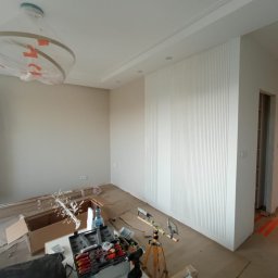 4domy - Pokój w trakcie wykończenia: białe pionowe panele ścienne, zakryta lampa, narzędzia i materiały budowlane na podłodze, widoczny fragment wykończonej łazienki w tle.