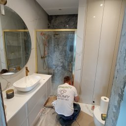 4domy - Pracownik w koszulce firmowej instaluje kabinę prysznicową z elementami w kolorze złotym w nowoczesnej łazience z umywalką nablatową i okrągłym lustrem.