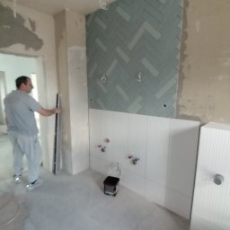 4domy - Wnętrze w trakcie prac wykończeniowych: ułożona niebieska mozaika w jodełkę na ścianie, częściowo wykończona biała ściana z instalacją wodną, pracownik z poziomicą w tle, widoczne ślady po...