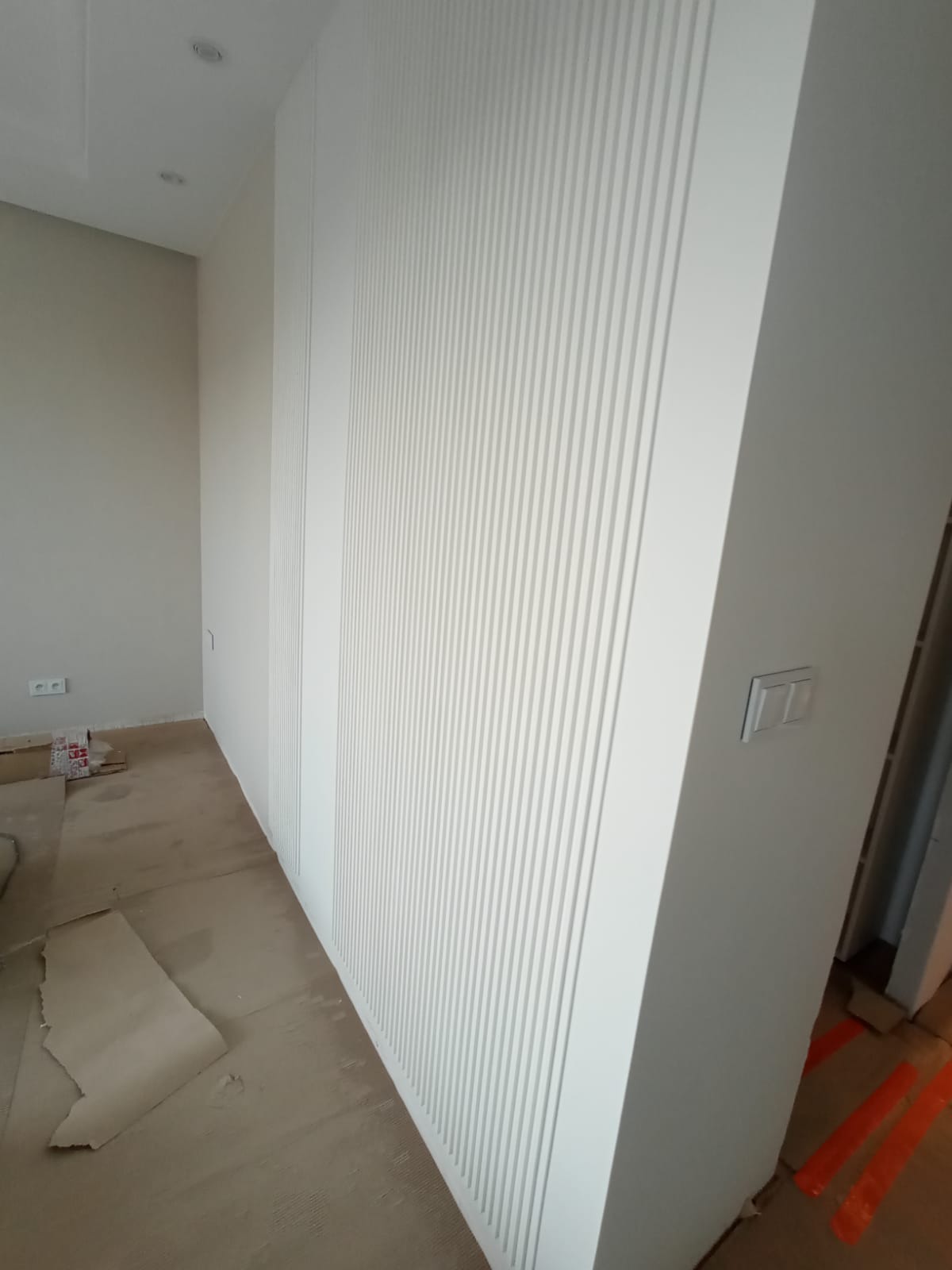 Biała ściana z dekoracyjnymi panelami pionowymi w trakcie montażu, widoczne gniazdko elektryczne, zabezpieczona podłoga i elementy konstrukcyjne w tle.