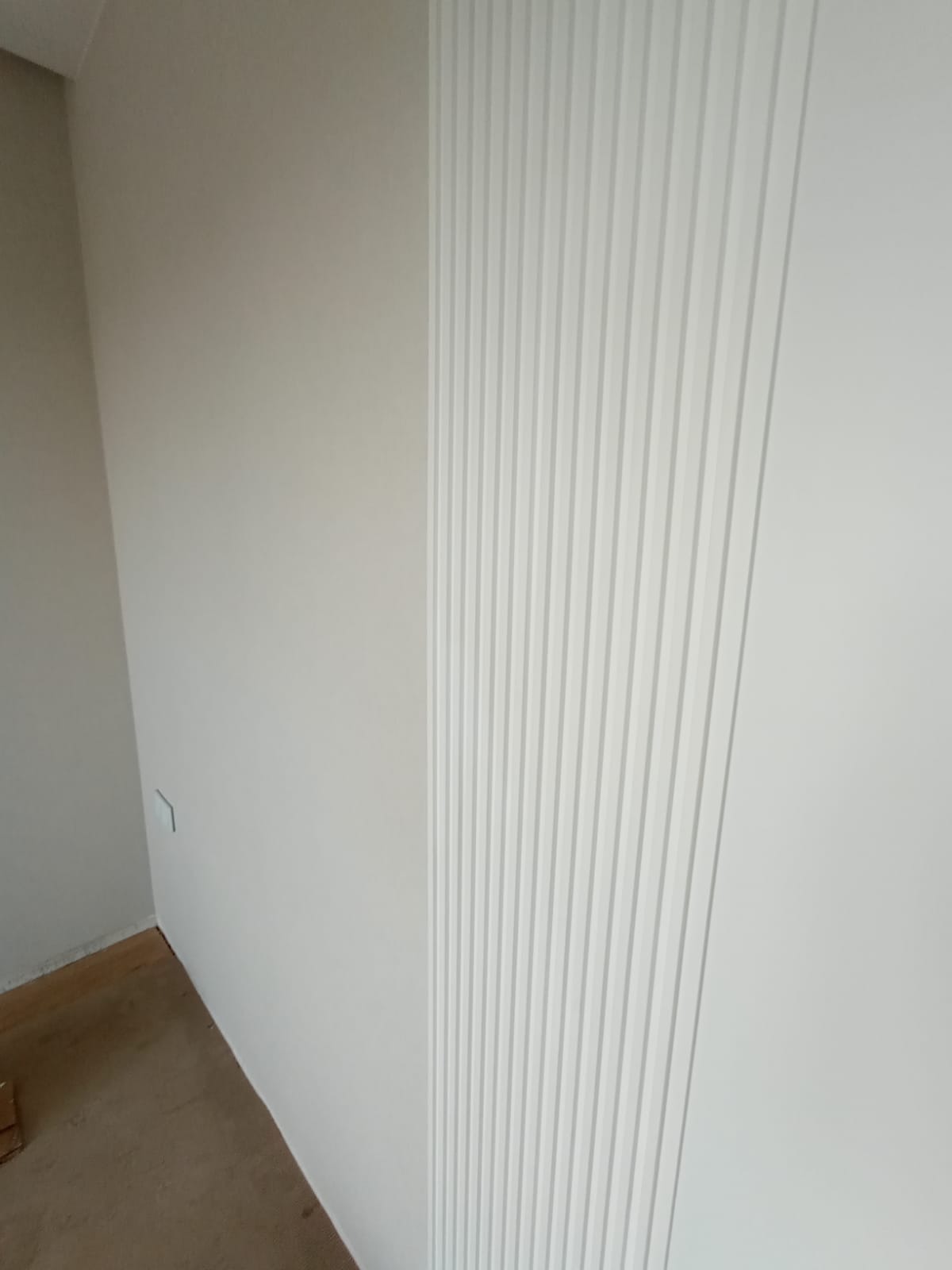 Biała ściana z dekoracyjnymi, pionowymi panelami ściennymi, widoczny fragment podłogi i ściana boczna w neutralnym odcieniu.