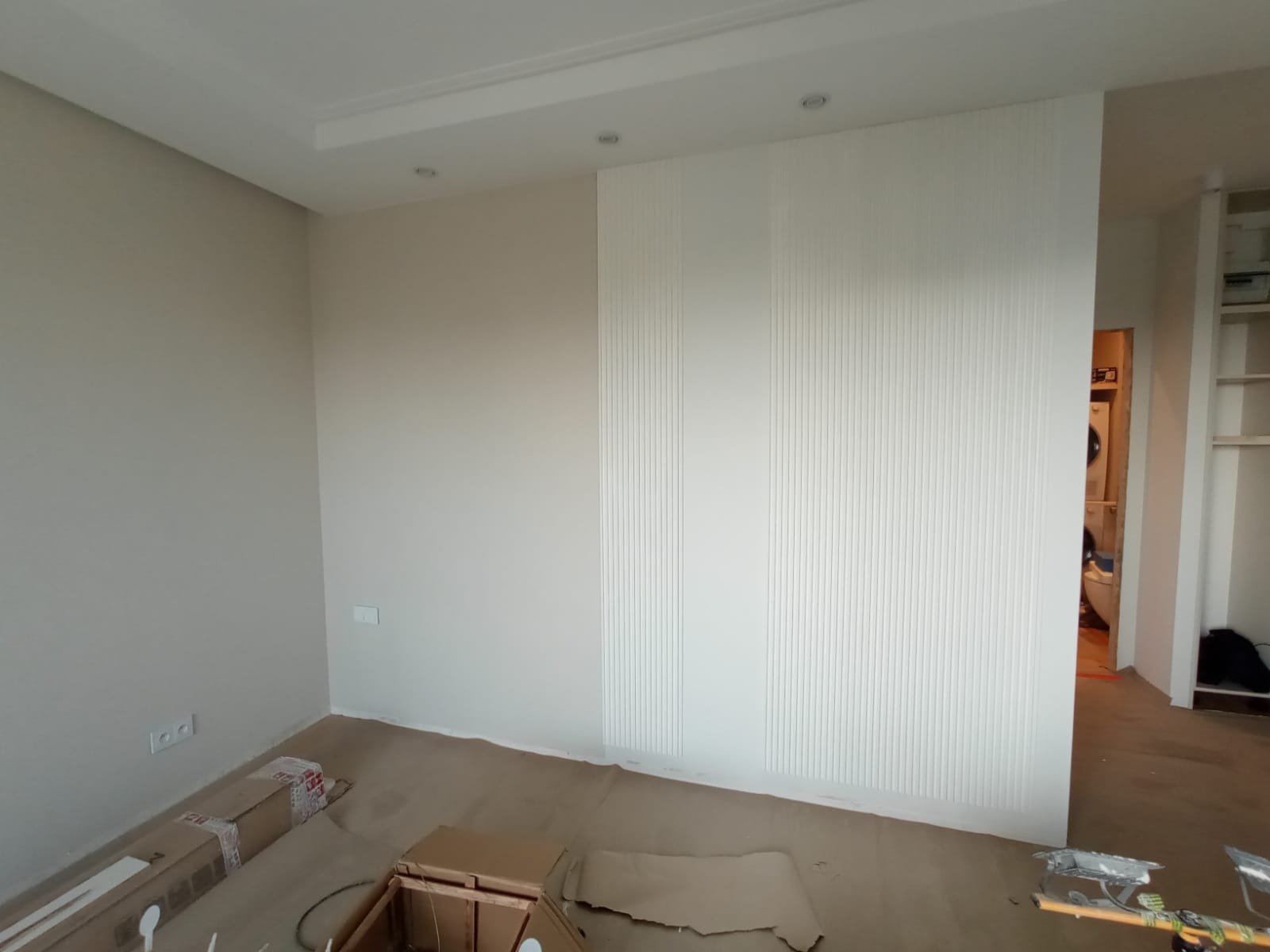 Pokój po odświeżeniu, z beżowymi ścianami, białą ścianą z pionowymi panelami dekoracyjnymi i widocznymi elementami kartonów na podłodze, sugerującymi prace wykończeniowe w toku.