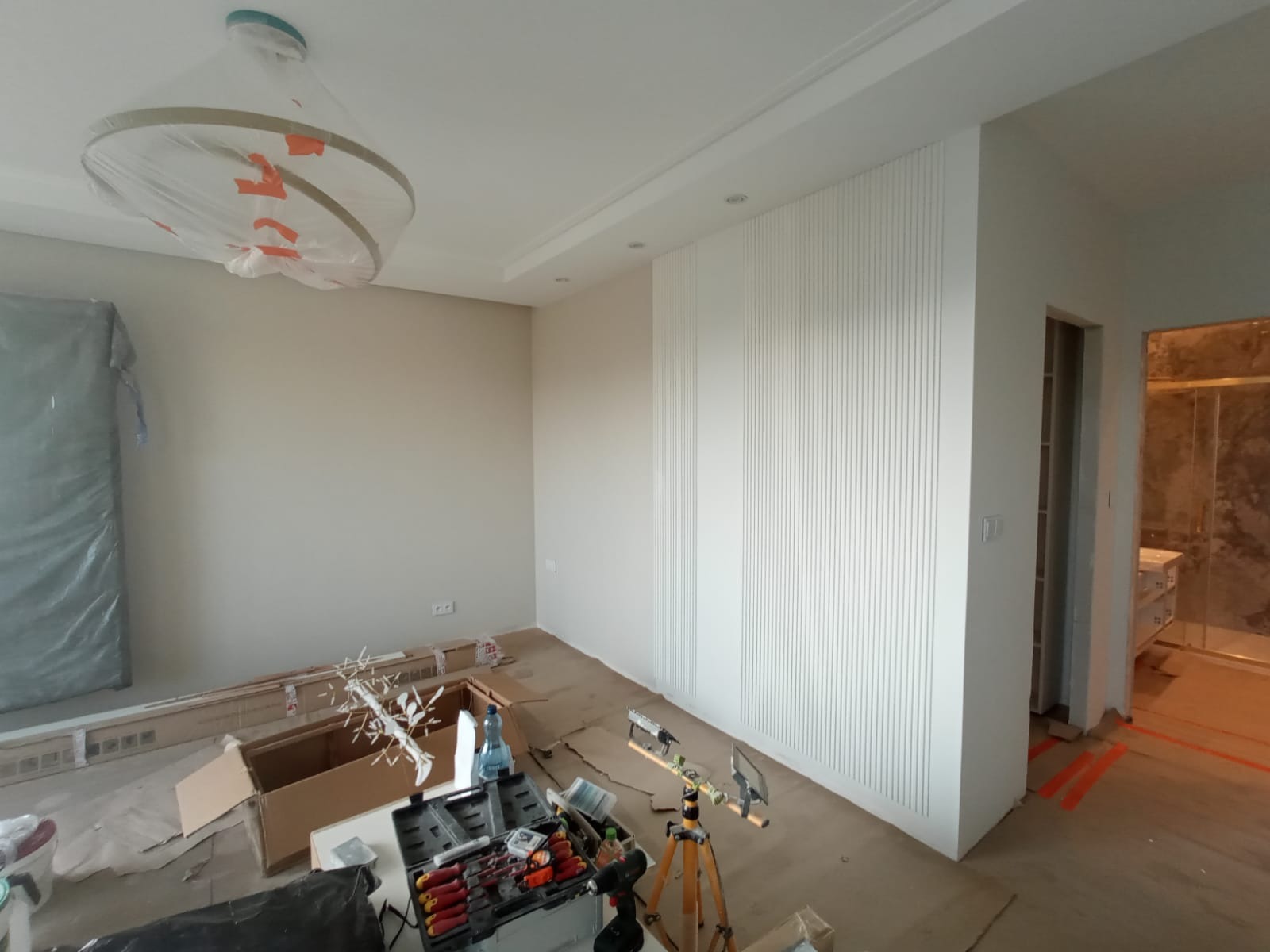Pokój w trakcie wykończenia: białe pionowe panele ścienne, zakryta lampa, narzędzia i materiały budowlane na podłodze, widoczny fragment wykończonej łazienki w tle.