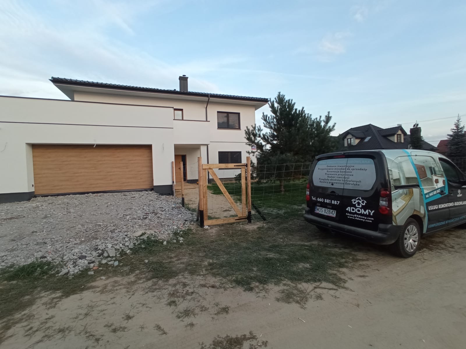 Nowoczesny dom jednorodzinny z brązową bramą garażową i drewnianym ogrodzeniem w trakcie montażu, obok zaparkowany samochód firmy remontowo-budowlanej z reklamą na karoserii.