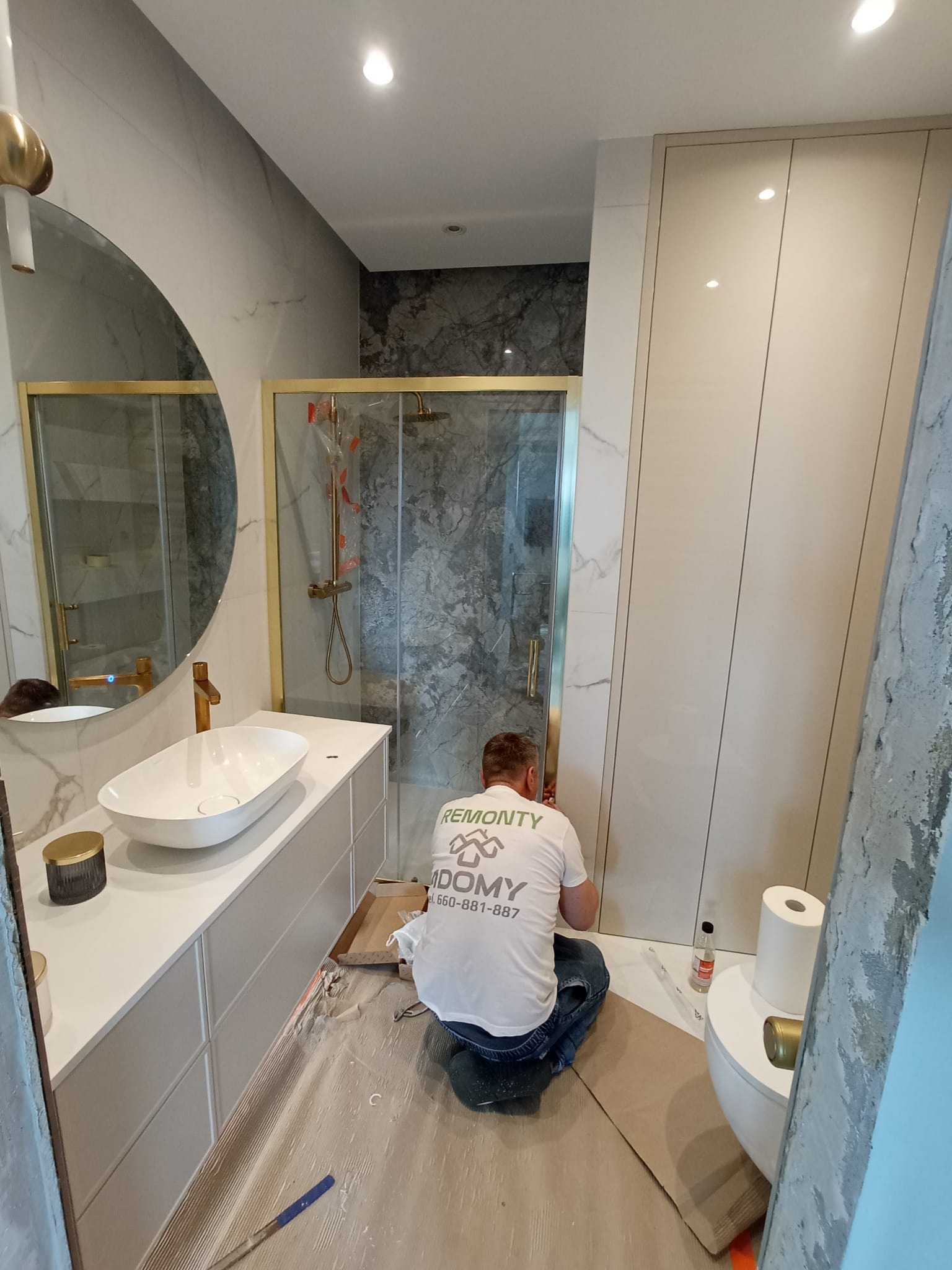 Pracownik w koszulce firmowej instaluje kabinę prysznicową z elementami w kolorze złotym w nowoczesnej łazience z umywalką nablatową i okrągłym lustrem.