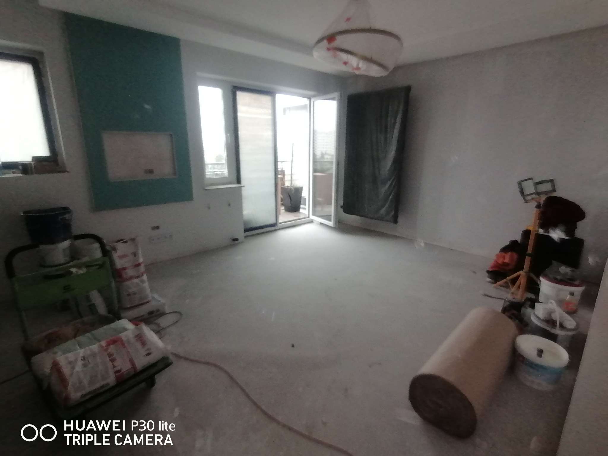 Wnętrze w trakcie prac przygotowawczych: surowe ściany po szpachlowaniu, podłoga z wylewką, widoczne materiały budowlane, narzędzia malarskie i wózek transportowy.