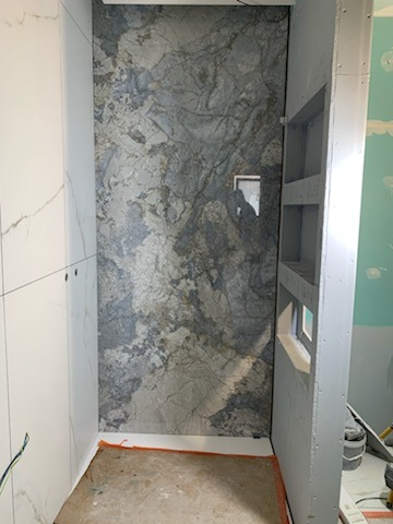 Wnęka prysznicowa w trakcie wykańczania, z płytkami imitującymi marmur na ścianie i wnęką z półkami po prawej stronie, widoczne niedociągnięcia w wykończeniu podłogi i łączeń.