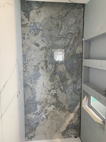 Płytka ścienna imitująca marmur w odcieniach szarości i błękitu, z kwadratowym odpływem liniowym umieszczonym centralnie, widoczna w narożniku łazienki z elementami konstrukcyjnymi półki wnękowej...