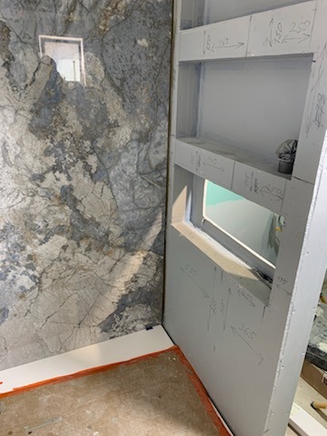 Ściana łazienki z płytami imitującymi marmur, sąsiadująca z konstrukcją z płyty karton-gips z wnękami, na której widoczne są odręczne notatki z wymiarami.