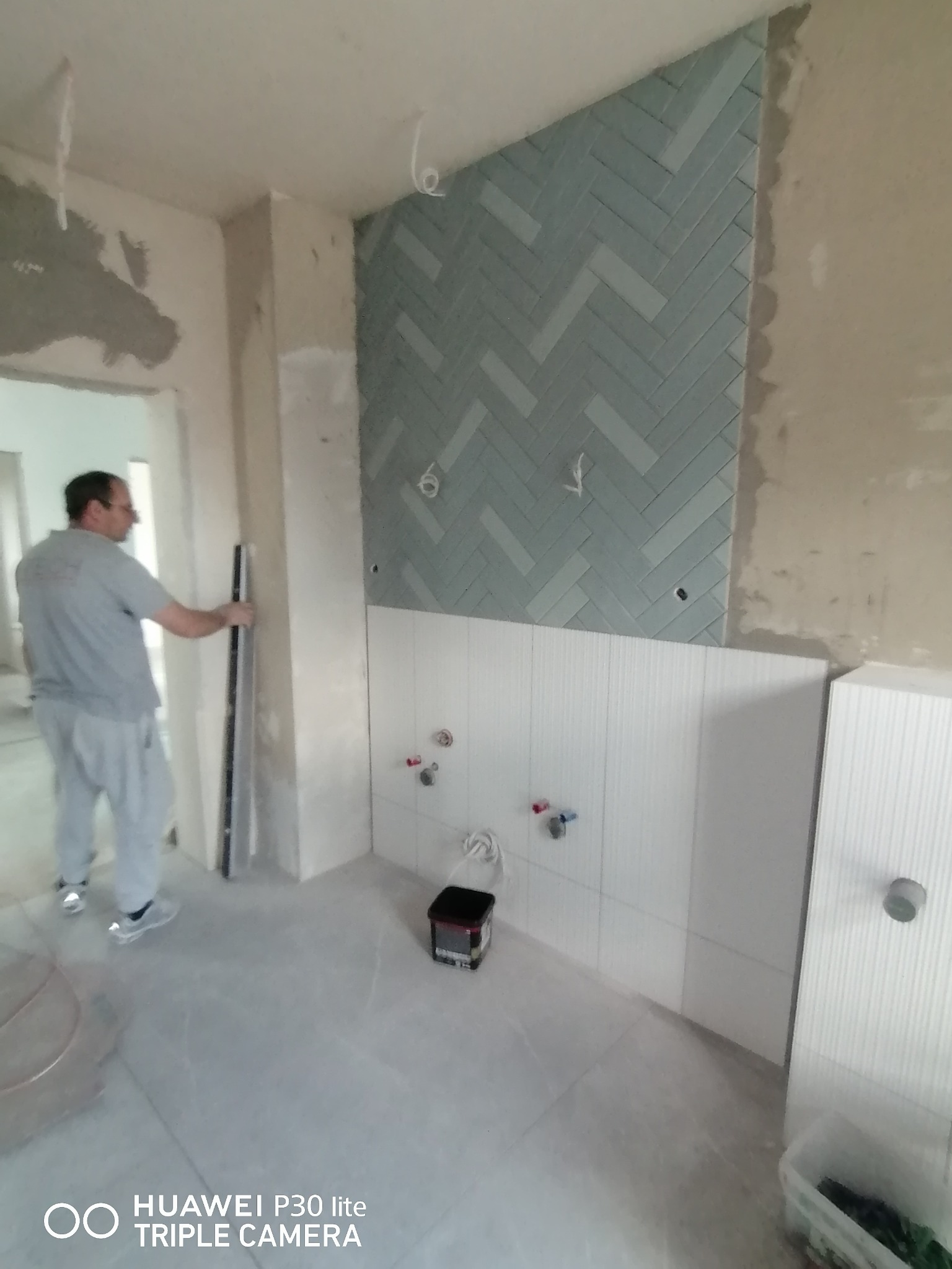 Wnętrze w trakcie prac wykończeniowych: ułożona niebieska mozaika w jodełkę na ścianie, częściowo wykończona biała ściana z instalacją wodną, pracownik z poziomicą w tle, widoczne ślady po...