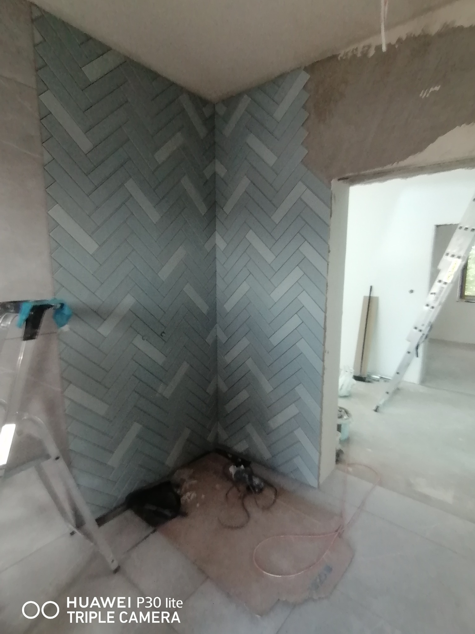 Ściana wyłożona jasnoszarą glazurą w jodełkę, widoczne narzędzia glazurnicze na podłodze i fragment drabiny w tle, surowy stan wykończenia pomieszczenia.