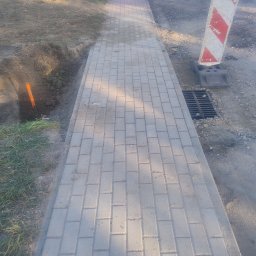 Ilona Witek - Ułożona szara kostka brukowa chodnika, obok wykop z pomarańczową rurą, w tle ziemia i czerwono-biały znak ostrzegawczy przy studzience kanalizacyjnej.