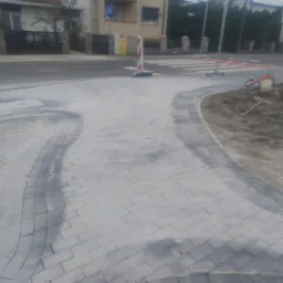 Nowo ułożona szara kostka brukowa na chodniku, z widocznymi śladami piasku i narzędziami budowlanymi w tle, obok ulicy z przejściem dla pieszych.