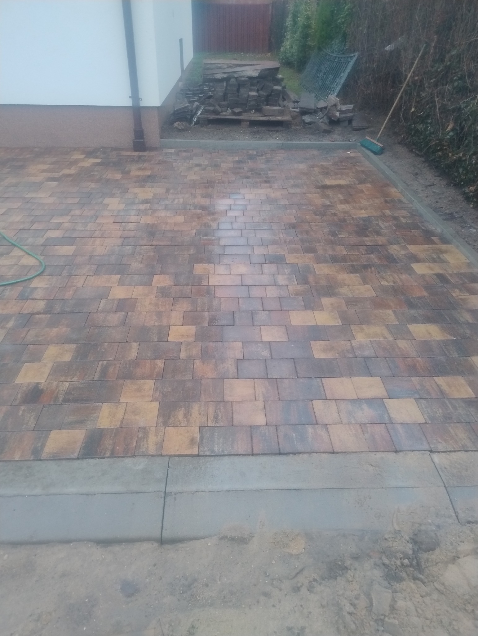 Ułożona kostka brukowa w odcieniach brązu i beżu tworząca wzór na podjeździe, z paletą kostki i narzędziami w tle, częściowo ukończona obrzeża.