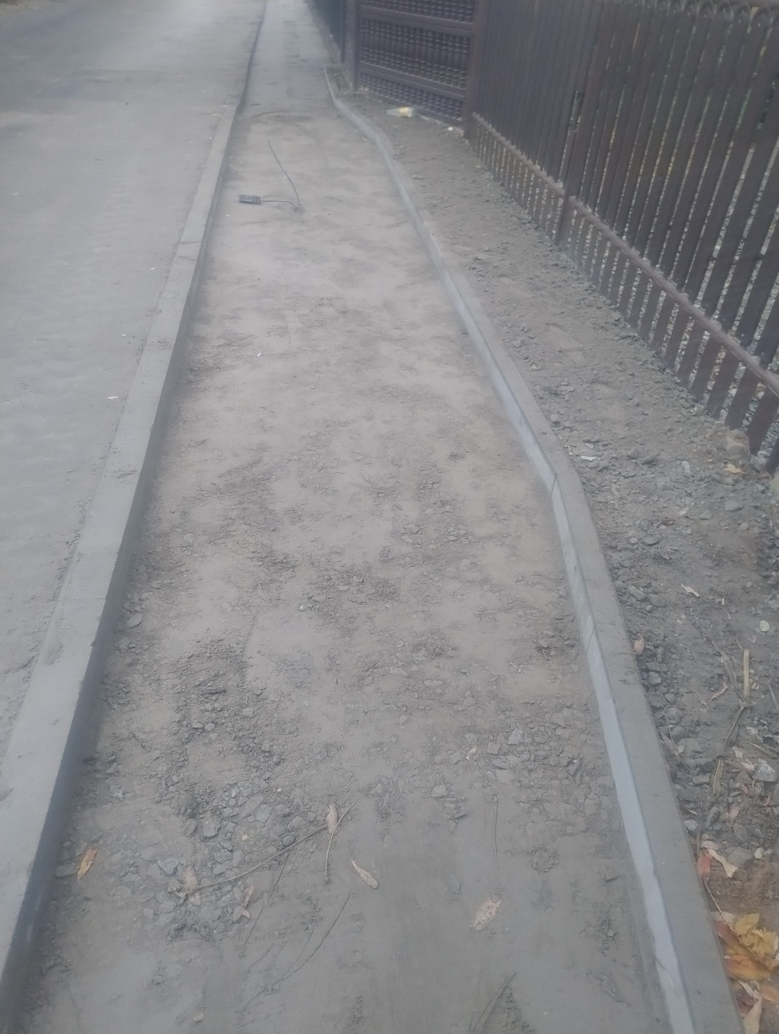 Przygotowanie podłoża pod chodnik z obrzeżami betonowymi, widoczne wyrównane podłoże z elementami kruszywa i liści, w tle metalowe ogrodzenie.