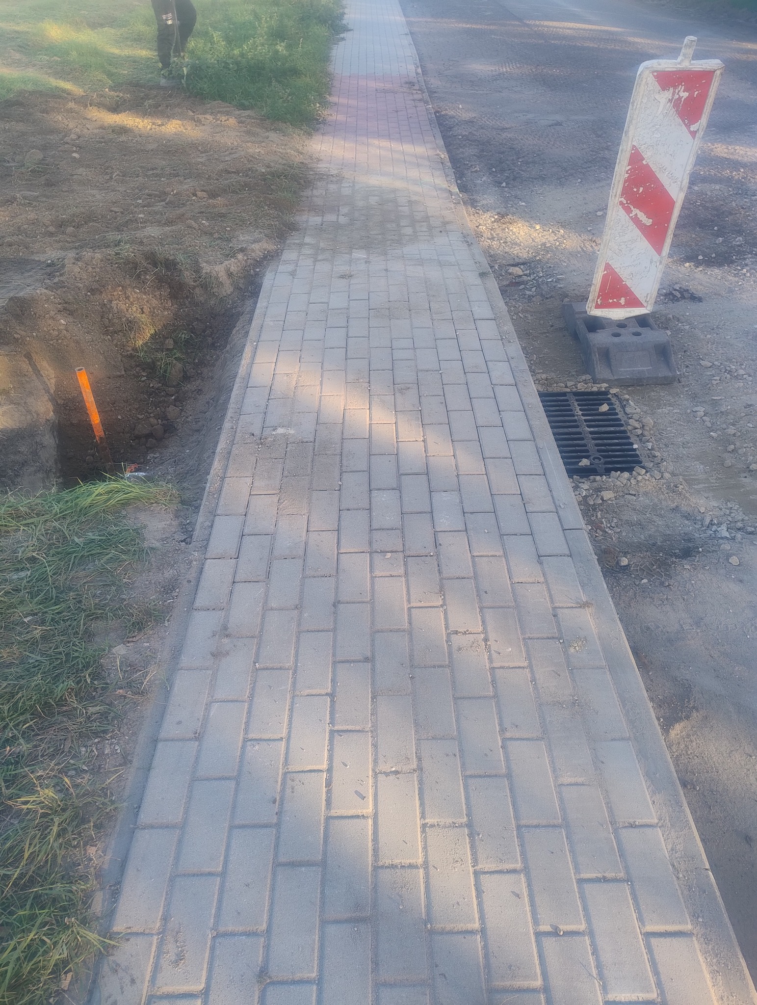 Ułożona szara kostka brukowa chodnika, obok wykop z pomarańczową rurą, w tle ziemia i czerwono-biały znak ostrzegawczy przy studzience kanalizacyjnej.