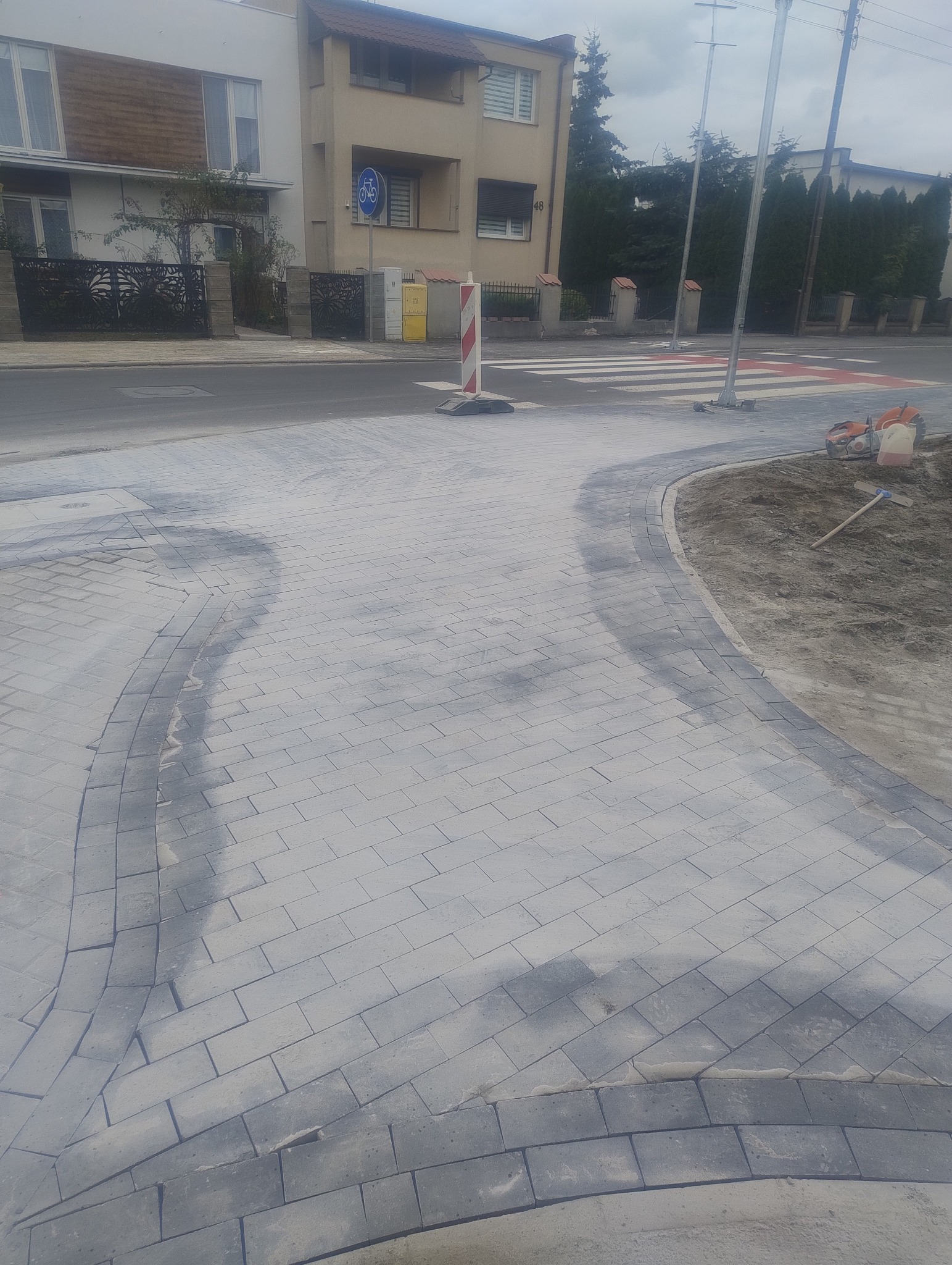 Nowo ułożona szara kostka brukowa na chodniku, z widocznymi śladami piasku i narzędziami budowlanymi w tle, obok ulicy z przejściem dla pieszych.