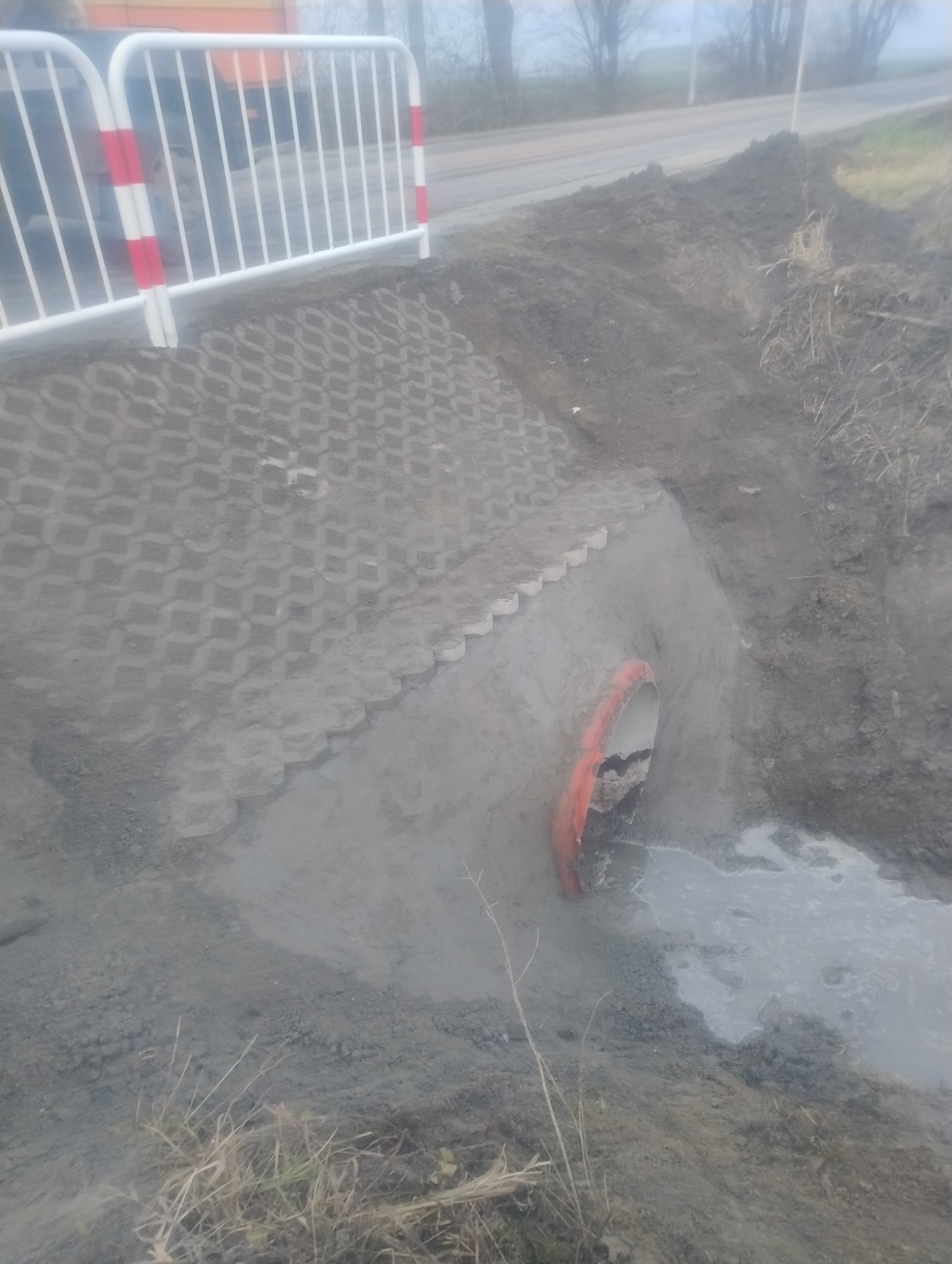 Ukończony drenaż z rurą odprowadzającą wodę, zabezpieczony ażurowymi płytami betonowymi i barierą ochronną przy drodze.